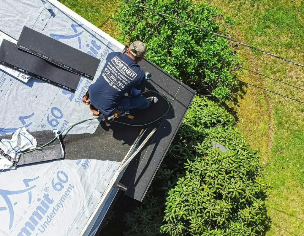 A worker installs roofing shingles on a house roof, wearing a safety harness and surrounded by greenery. Roofing materials and underlayment are also visible.