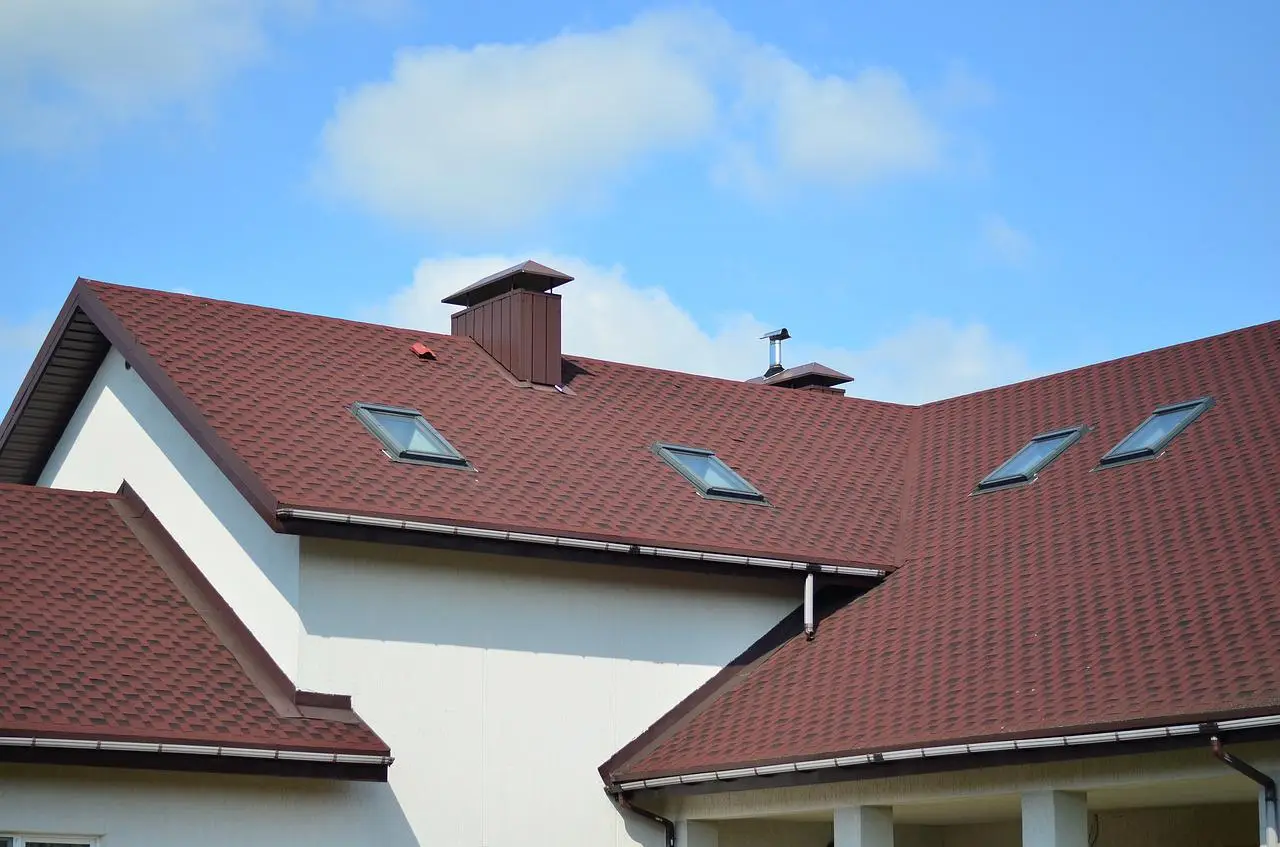 A modern house with a red shingled roof featuring several skylights and a chimney against a blue sky with scattered clouds.