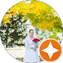 A bride in a white dress holds a red bouquet while standing outdoors under trees with yellow autumn leaves.