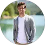 A man wearing a white t-shirt and gray cardigan stands outdoors near a body of water, with trees and hills in the background.