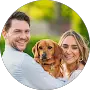 A man and a woman outdoors smiling at the camera, holding a brown dog between them, with green foliage in the background.